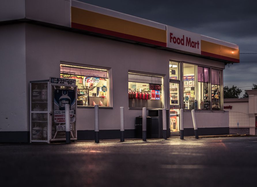 The image depicts the exterior of a Food Mart convenience store during dusk, illuminated by interior lighting that highlights the shop's large glass windows. Reflected in the windows are shelves stock