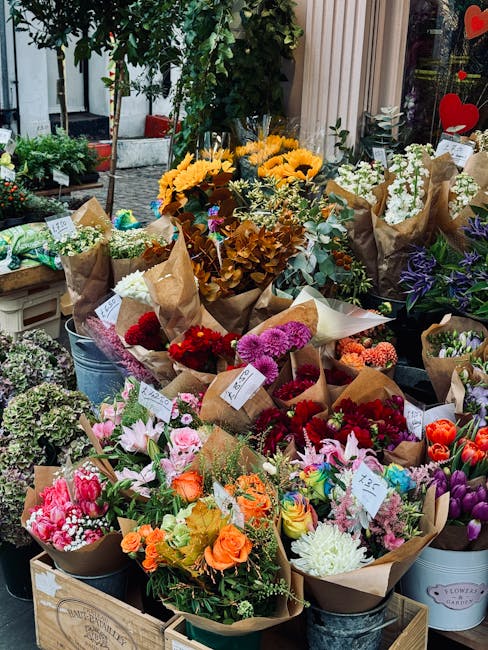 An outdoor display of various fresh flower bouquets arranged on wooden crates and metal buckets, featuring an array of vibrant colors including reds, pinks, oranges, whites, and purples. The bouquets 
