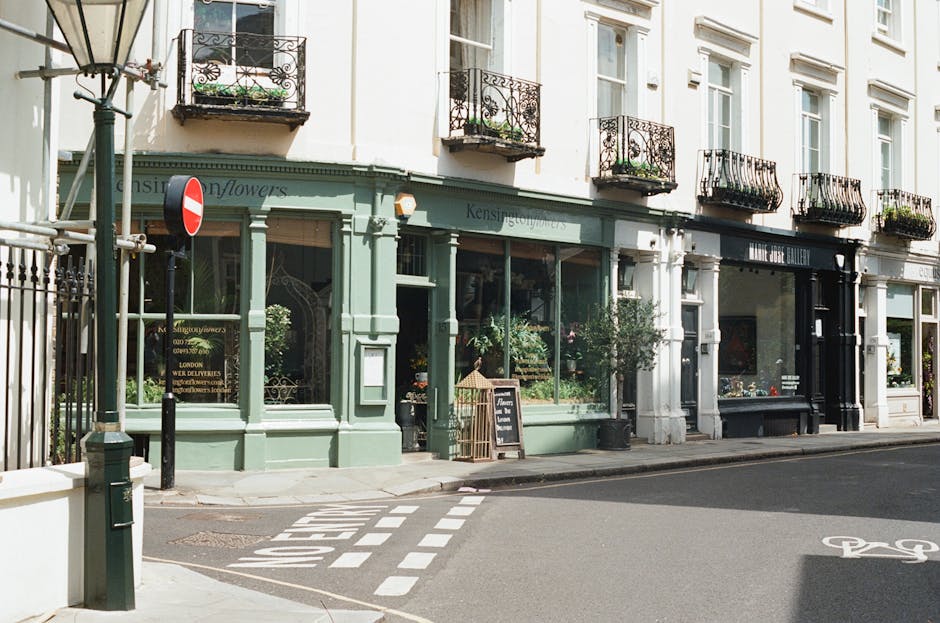 A street view featuring a vintage-style flower shop with a pale green facade and large glass windows displaying floral arrangements inside. The shop has a black sign above the window, and a small chal